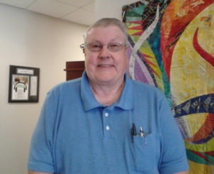 Carl Babb, volunteer at AINC, standing in the AINC lobby smiling. blue shirt with glasses.