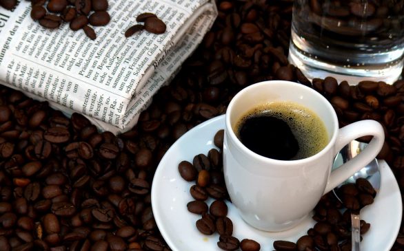 coffee sitting on a small plate covered in coffee beans. coffee beans cover table and a newspaper is visible on the table.