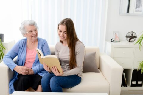 A young woman reading to an older woman on a couch in a sunny room.
