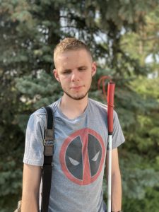 Evan standing in front of trees with a white cane with a red handle. He is wearing a graphic Tshirt and carrying a bag.