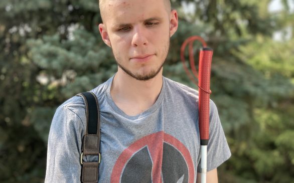 Evan standing in front of trees with a white cane with a red handle. He is wearing a graphic Tshirt and carrying a bag.