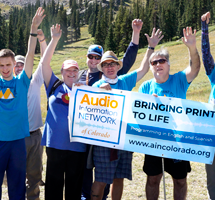 The AINC Hiking team, the AINC Audio Trekkers, standing and cheering in a mountain scene scape holding an AINC Bringing print to Life banner.