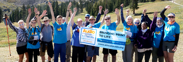 The AINC Hiking team, the AINC Audio Trekkers, standing and cheering in a mountain scene scape holding an AINC Bringing print to Life banner.