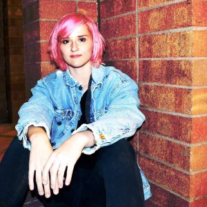 Alex sitting infront of a brick wall with a short pink haircut and a denim jacket