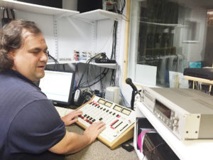 Ryan sitting in the old control room with his hands on the audio board, smilling and looking back at the camera.