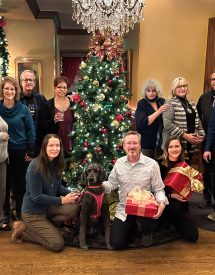AINC staff and board posing around a christmas tree, smiling. Some people are holding gift wrapped presents, and a poodle guide dog sits in the front.