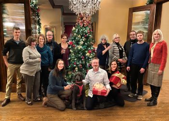 AINC staff and board posing around a christmas tree, smiling. Some people are holding gift wrapped presents, and a poodle guide dog sits in the front.