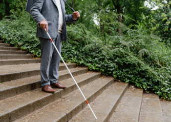 Man walking down outdoor staircase wearing a suite, holding a white cane and an umbrella