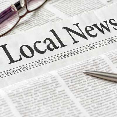 closeup image of a newspaper that says "local news" with glasses and pen.